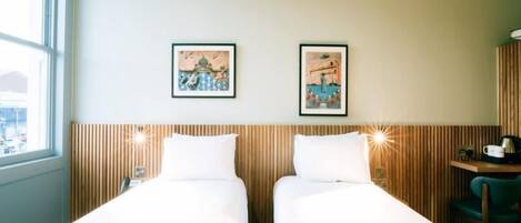 Une chambre d’hôtel avec deux lits simples, une tête de lit en bois, une petite table avec une chaise et deux tableaux encadrés au mur.