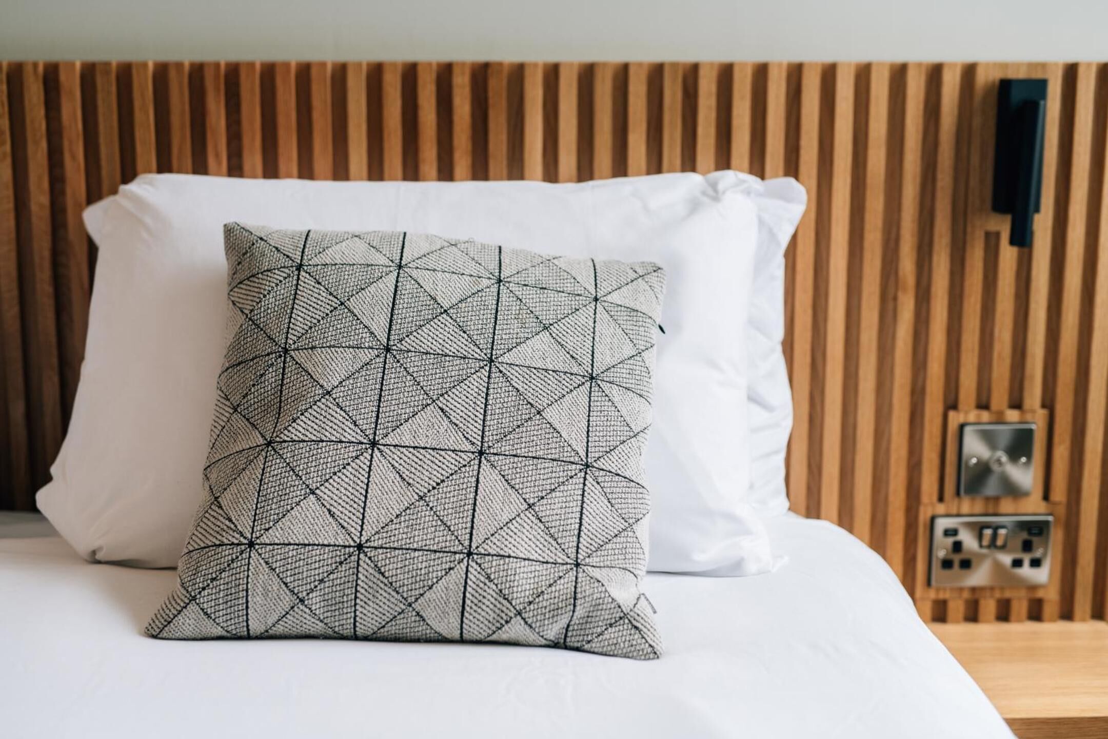Een bed met een sierkussen met een geometrisch patroon en een effen wit kussen, voor een houten hoofdbord.