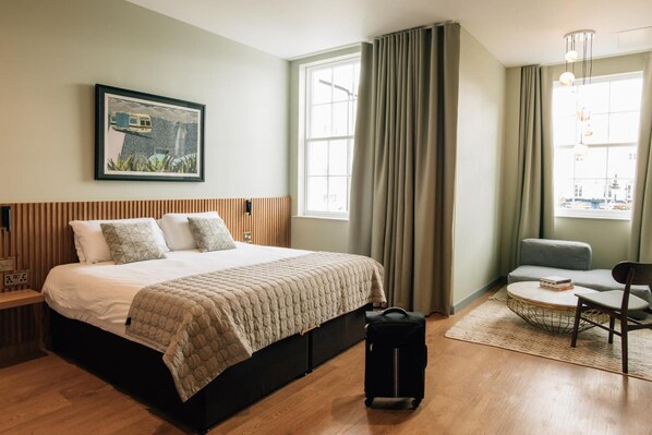 A hotel room with a large bed, a desk, a chair, a window with curtains, and a painting on the wall.