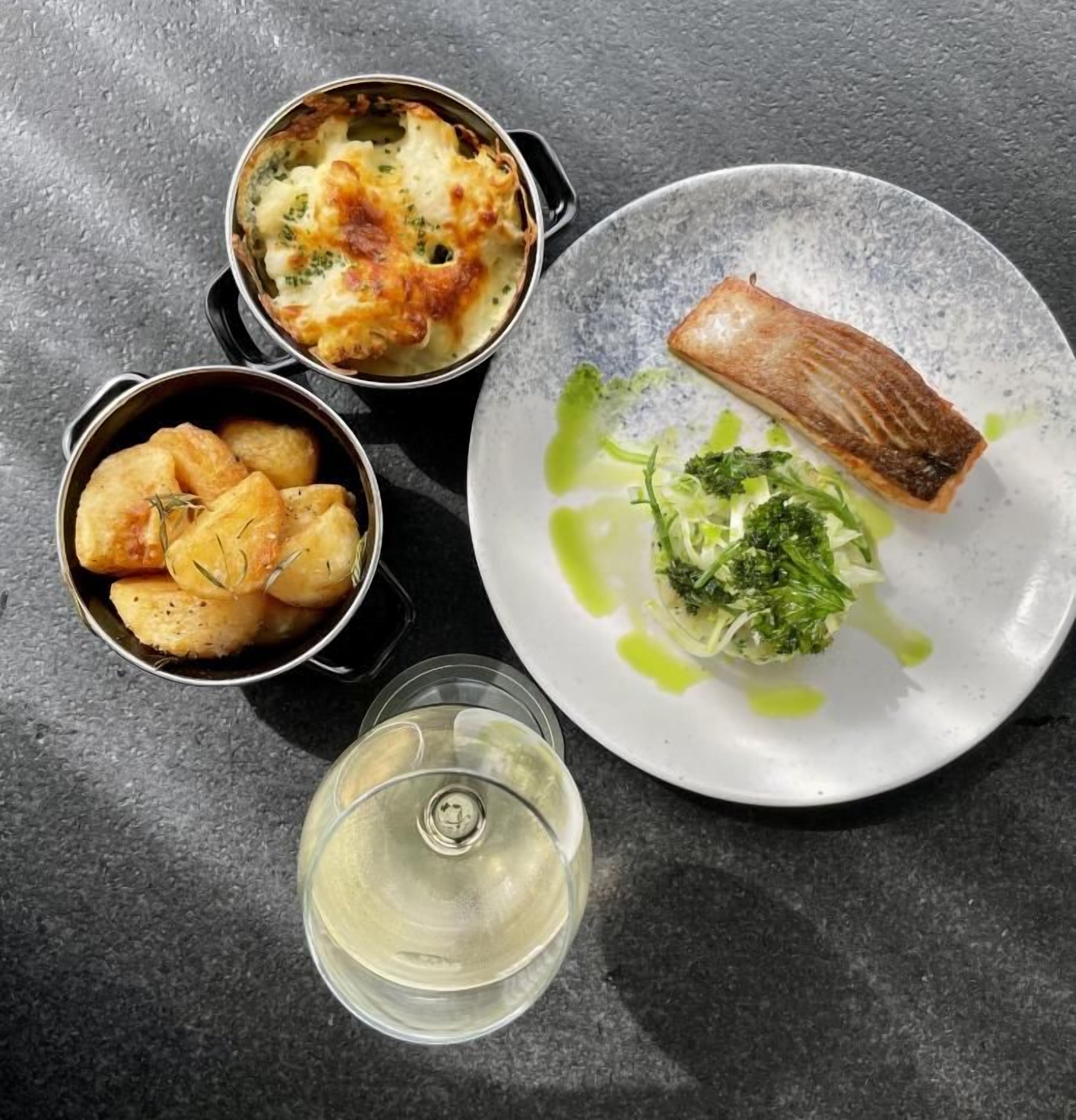 Un plato de salmón, un bol de patatas y una copa de vino blanco sobre una superficie oscura.