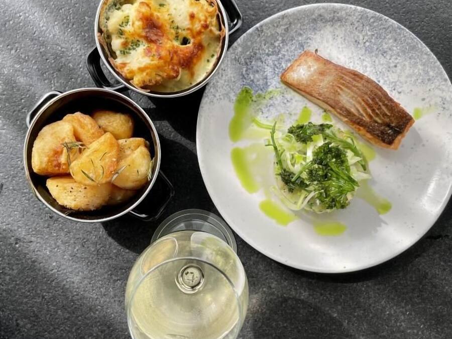 Ein Teller mit Lachs, eine Schüssel Kartoffeln und ein Glas Weißwein auf einer dunklen Oberfläche.