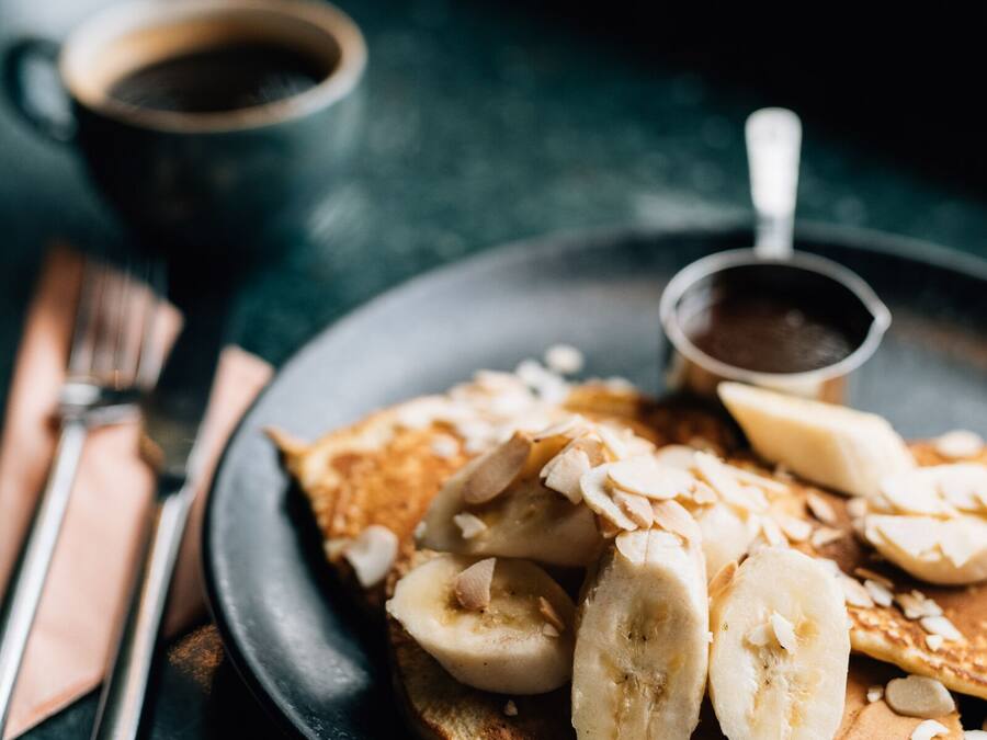 Une assiette de crêpes garnie de tranches de bananes et d’amandes, accompagnée d’un petit bol de sirop, le tout posé sur une surface sombre, avec une fourchette et un couteau à côté.
