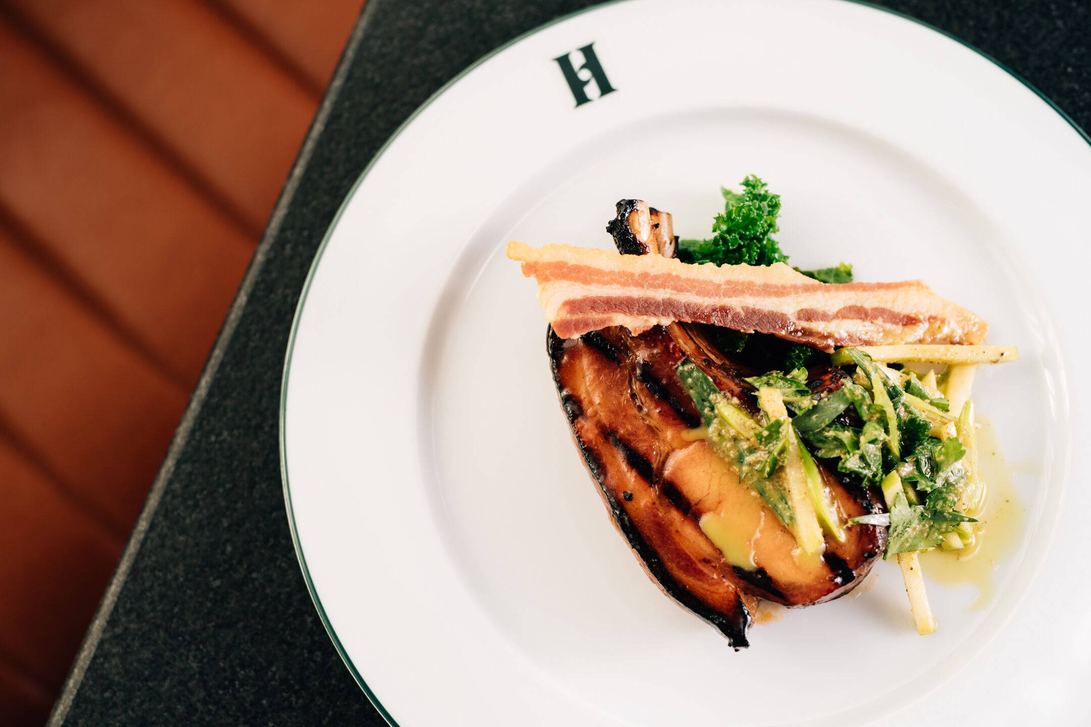 Prato com carne grelhada, bacon e vegetais em um prato branco com a letra 'H'.