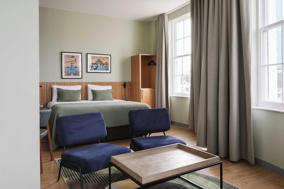 A hotel room with a bed, two blue armchairs, a small table, a wooden wardrobe, and two framed pictures on the wall.
