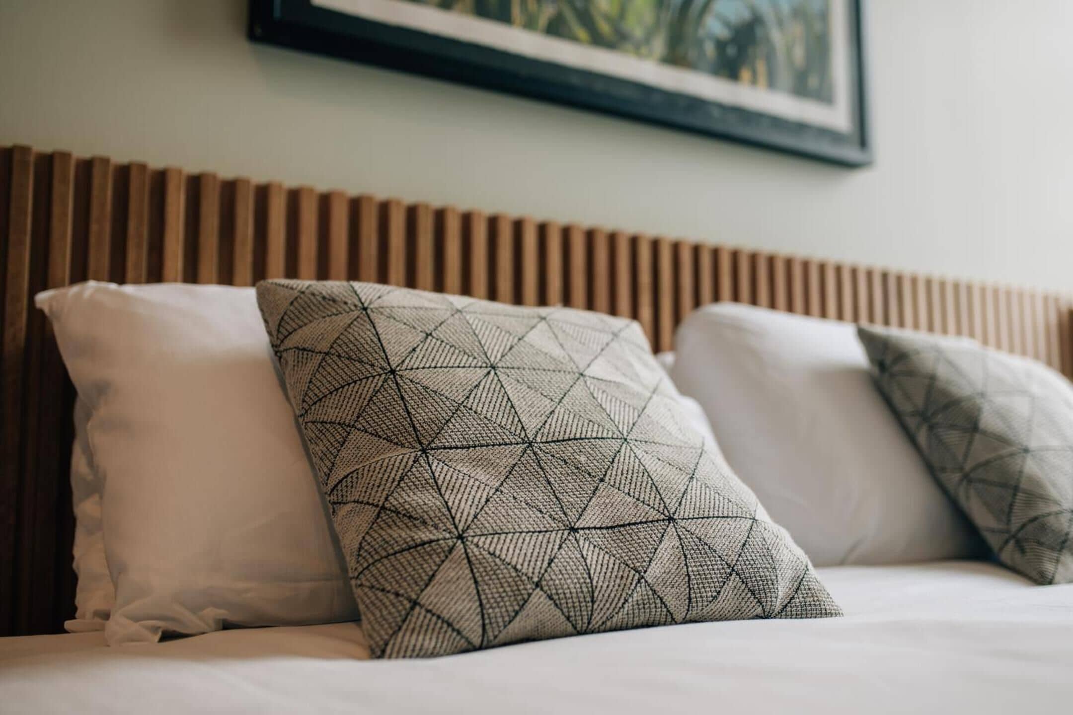 Una cama con tres almohadas, una blanca lisa y dos con diseños geométricos, frente a un cabecero de madera.