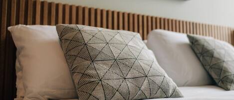 Una cama con tres almohadas, una blanca lisa y dos con diseños geométricos, frente a un cabecero de madera.