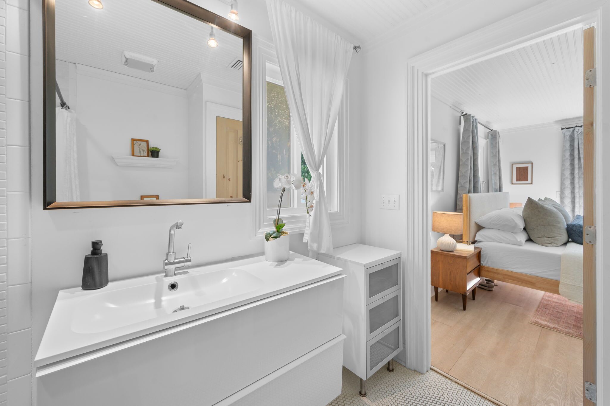 Un baño moderno con un espejo grande, lavamanos blanco y una mesita de noche pequeña con lámpara.