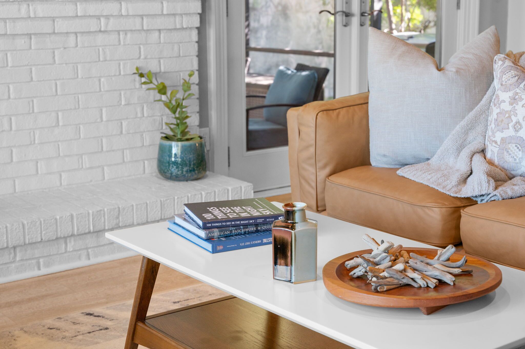 A modern living room with a white brick wall, a leather sofa, a white coffee table with books and a bottle, and a wooden tray with decorative items.