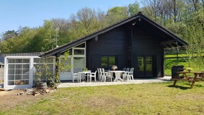 Una cabaña de madera con un patio que incluye una mesa, sillas, una parrilla y un cobertizo pequeño.