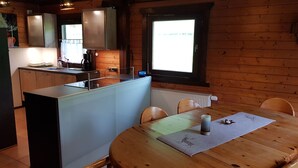 Una cocina acogedora de estilo cabaña con paredes de madera, una mesa de comedor con sillas, una isla integrada con un fregadero y un ventanal que proporciona luz natural.