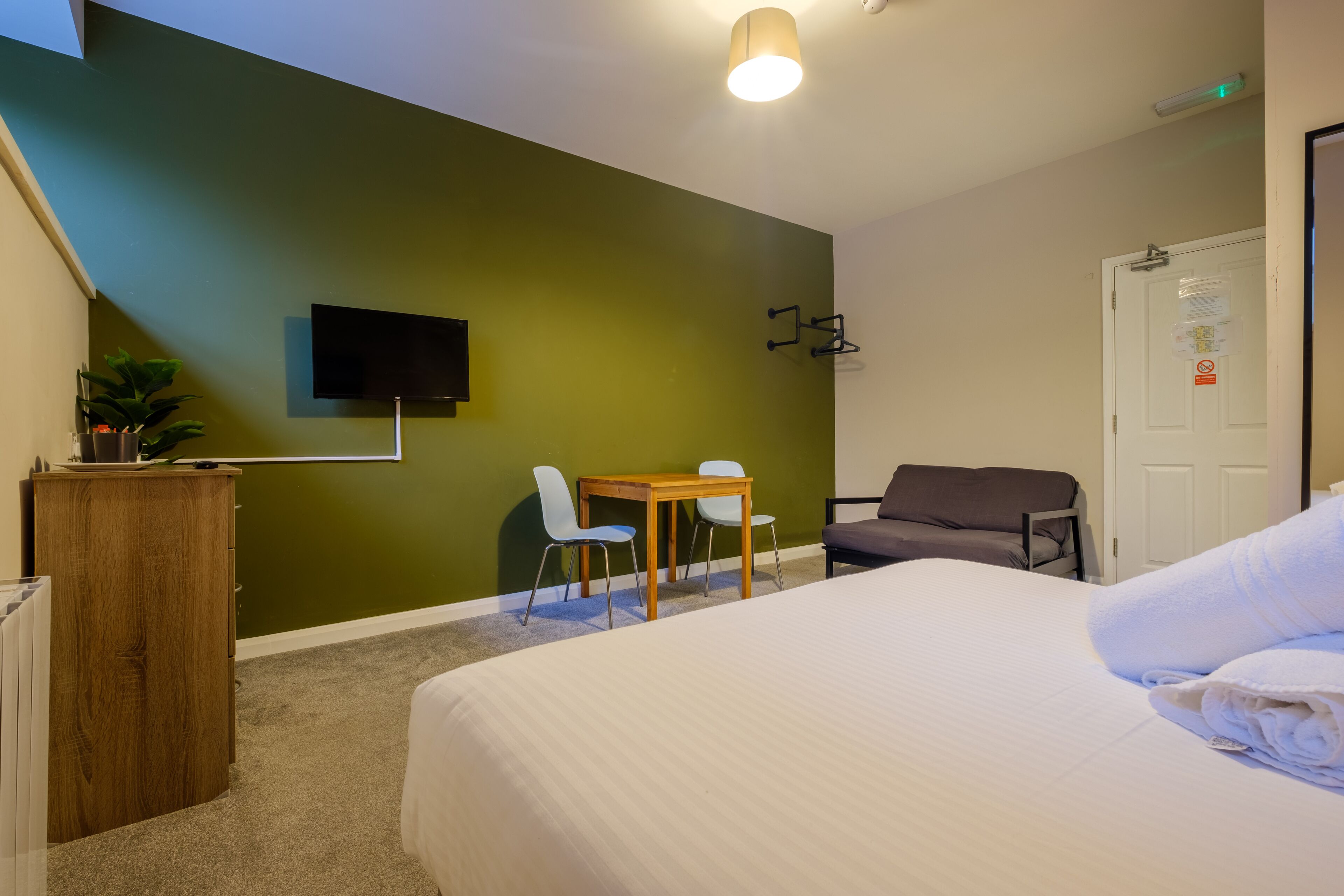 Quarto de hotel com cama, escrivaninha, duas cadeiras, televisão e sofá.