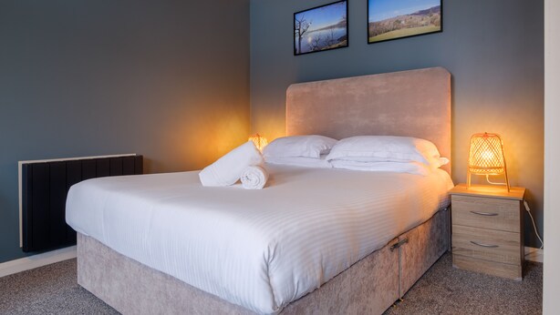 A neatly made bed with white linens and a rolled towel, a bedside table with a lamp, and two framed pictures on the wall. - Station Lodge (Windermere)