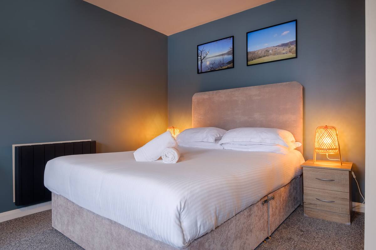 Un lit bien fait, avec du linge de lit blanc et une serviette pliée, une table de chevet avec une lampe, et deux tableaux encadrés accrochés au mur.