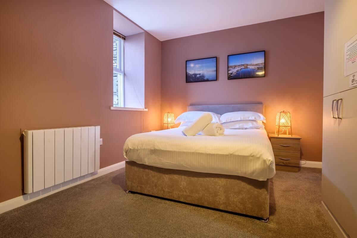 Une chambre à coucher avec un grand lit, deux lampes de chevet, une fenêtre et deux tableaux encadrés au mur.