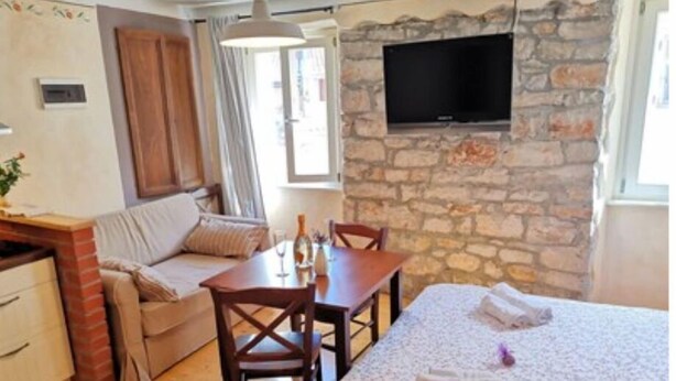 Ein gemütliches Zimmer mit Steinwand, einem flachbildfernseher über einem Holztisch, einer Couch mit Kissen und einem Bett mit blumigem Überwurf.