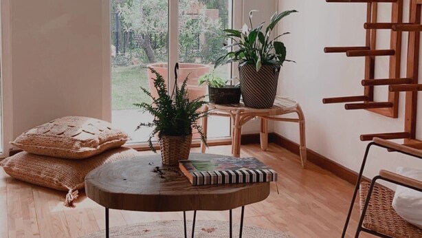 Ein geräumiges Zimmer mit großem Fenster, Holzboden, rundem Tisch mit Teppich und einem Holzregal an der Wand.