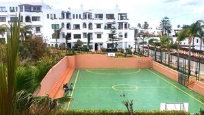Un terrain de basket en plein air, entouré d’une végétation luxuriante, et un bâtiment blanc doté de nombreux balcons.