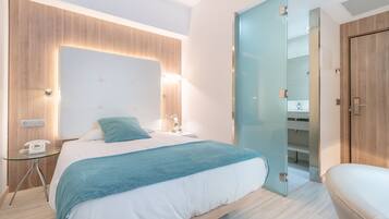 A modern hotel room with a large bed, bedside table, and a glass partition separating the bedroom from the bathroom.