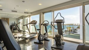 Uma academia com máquinas de cardio, um remo e vista para o mar através de amplas janelas.
