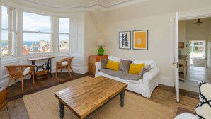 Ein geräumiges Wohnzimmer mit großem Fenster, einem Holztisch, einem weißen Sofa mit gelben Kissen und einem Korbstuhl.