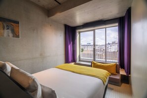 Habitación de hotel con cama, ventana con cortinas, vista de la ciudad y un cuadro en la pared.