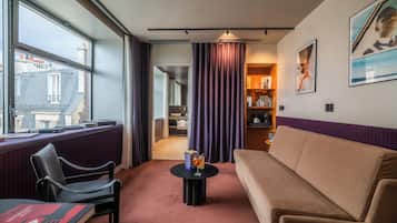 Une chambre d’hôtel avec un canapé, une petite table avec une boisson, une chaise, une fenêtre donnant sur des immeubles et une porte menant à un couloir.