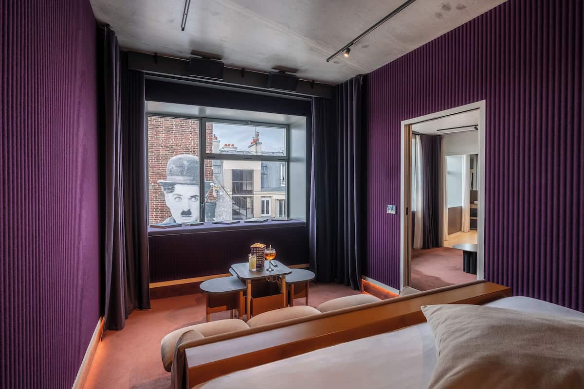 Une chambre d’hôtel avec une grande fenêtre, un lit, une petite table avec une boisson et un miroir.