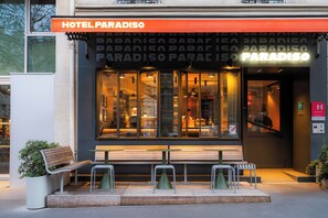 Entrada de un hotel con un cartel que dice 'HOTEL PARADISO', un banco afuera y una escalera que lleva a la entrada.