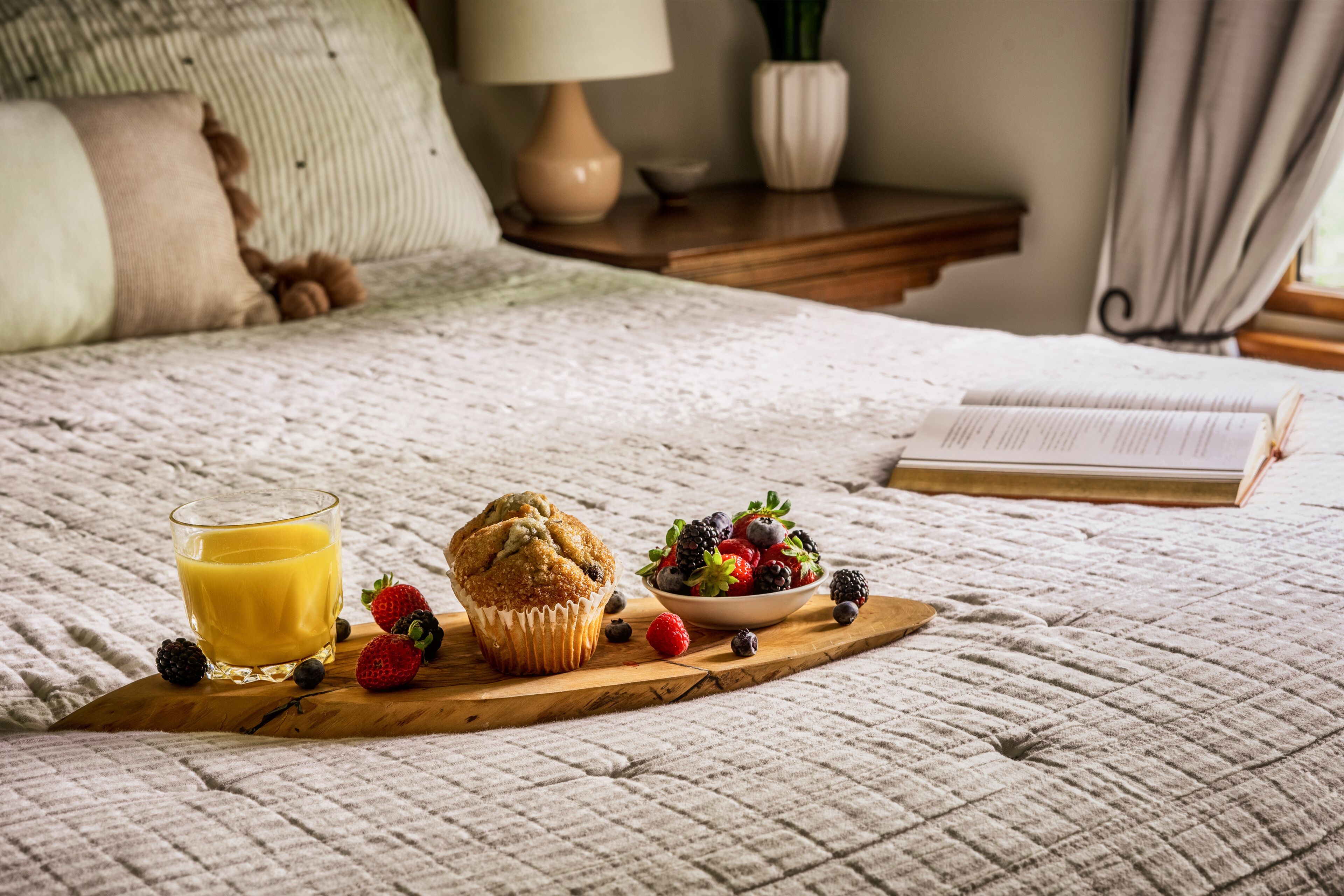 A bed with a quilted blanket, a wooden bedside table with a lamp, a bowl of fruit, a muffin, and a glass of orange juice on a tray.