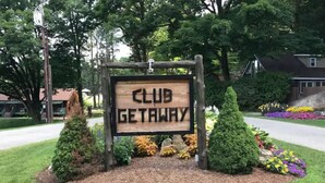 En träskylt med texten ”CLUB GETAWAY”, omgiven av vackert anlagda buskar och blommor.