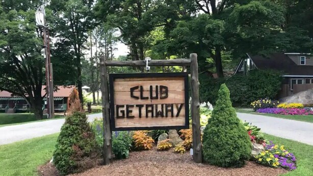 Un letrero de madera con la inscripción 'CLUB GETAWAY' rodeado de arbustos y flores cuidadosamente plantados.