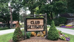 Uma placa de madeira com a inscrição 'CLUB GETAWAY', rodeada por arbustos e flores bem cuidados.