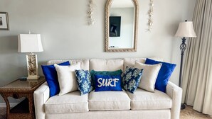 Un canapé beige agrémenté de coussins à motifs bleus et blancs, une petite table d’appoint avec une lampe, un miroir et un élément décoratif fixé au mur.