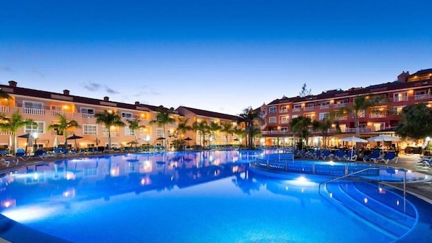 Una piscina amplia e iluminada rodeada de edificios de hotel con balcones y palmeras.