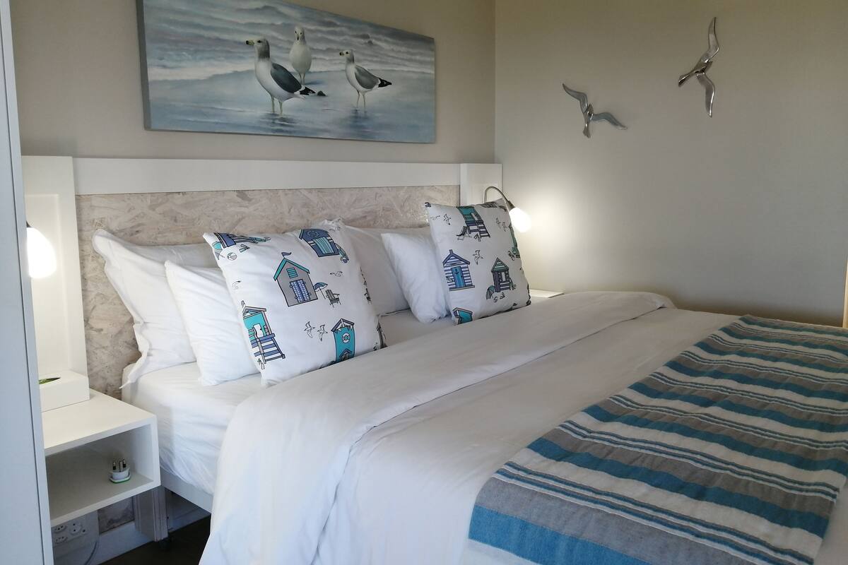 A neatly made bed with white bedding and patterned pillows, a painting of seagulls on the wall, and a striped blanket on the bed.