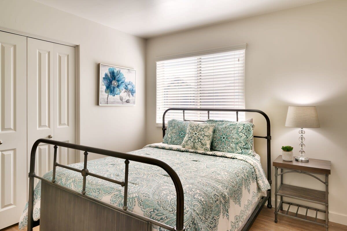A bedroom with a bed, a bedside table with a lamp, a window with blinds, and a door.
