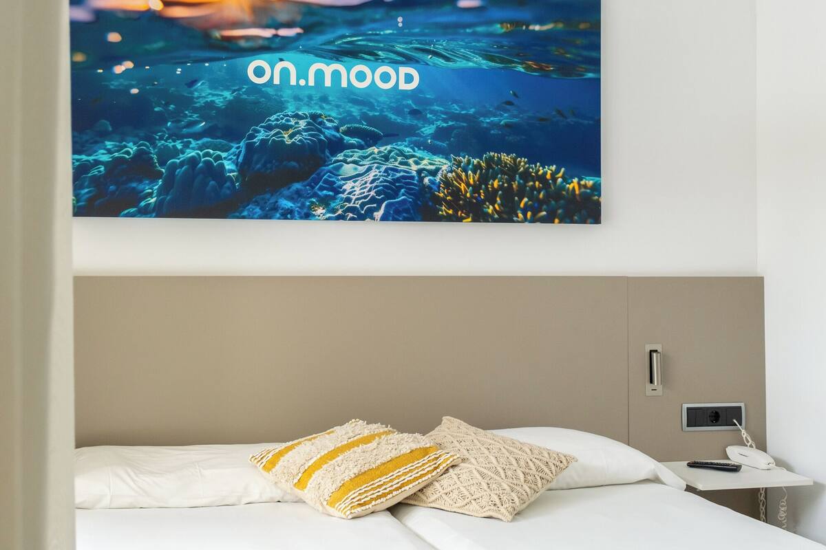 A hotel room with a bed, a nightstand, and a wall-mounted TV displaying a scenic underwater image with the text "on.mood".