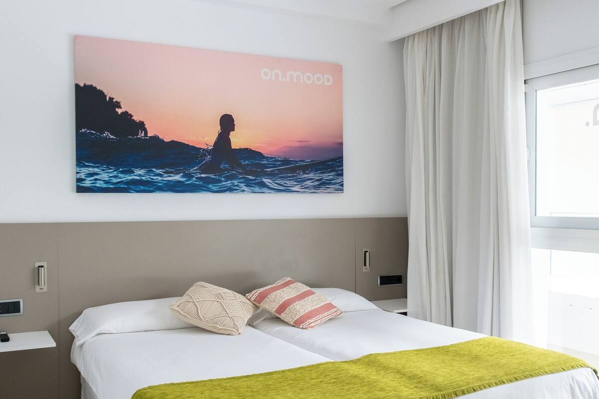 A modern hotel room with a large bed, a nightstand, a bedside table, a window with curtains, and a wall-mounted picture of a sunset over the ocean.