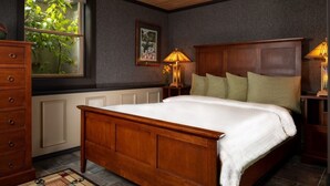 A bedroom with a wooden bed, two bedside lamps, a dresser, a window with a view of greenery, and a framed picture on the wall. - Riverside Chalet Plus (Confluence)