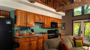 A kitchen with wooden cabinets, a black refrigerator, and a microwave. There is a dining table with a loaf of bread and a fruit bowl. A beige sofa with colorful cushions is in the foreground. - Riverside Chalet Plus (Confluence)