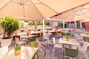 Een terras buiten met meerdere tafels en stoelen, waarvan sommigen onder parasols staan. Er staan potplanten en de vloer is van steen.