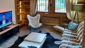 Interior acogedor de una cabaña de troncos con techo de madera, televisor, sillón blanco y mullido, sofá y alfombra de cuero de vaca.
