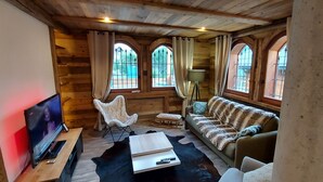 Interior de una cabaña de troncos con una televisión, una silla blanca, un sofá y ventanas amplias.