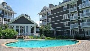 Ein Schwimmbad, umgeben von einer Pflasterterrasse, mit einem Gebäude im Hintergrund, das Balkone und einen überdachten Bereich aufweist.