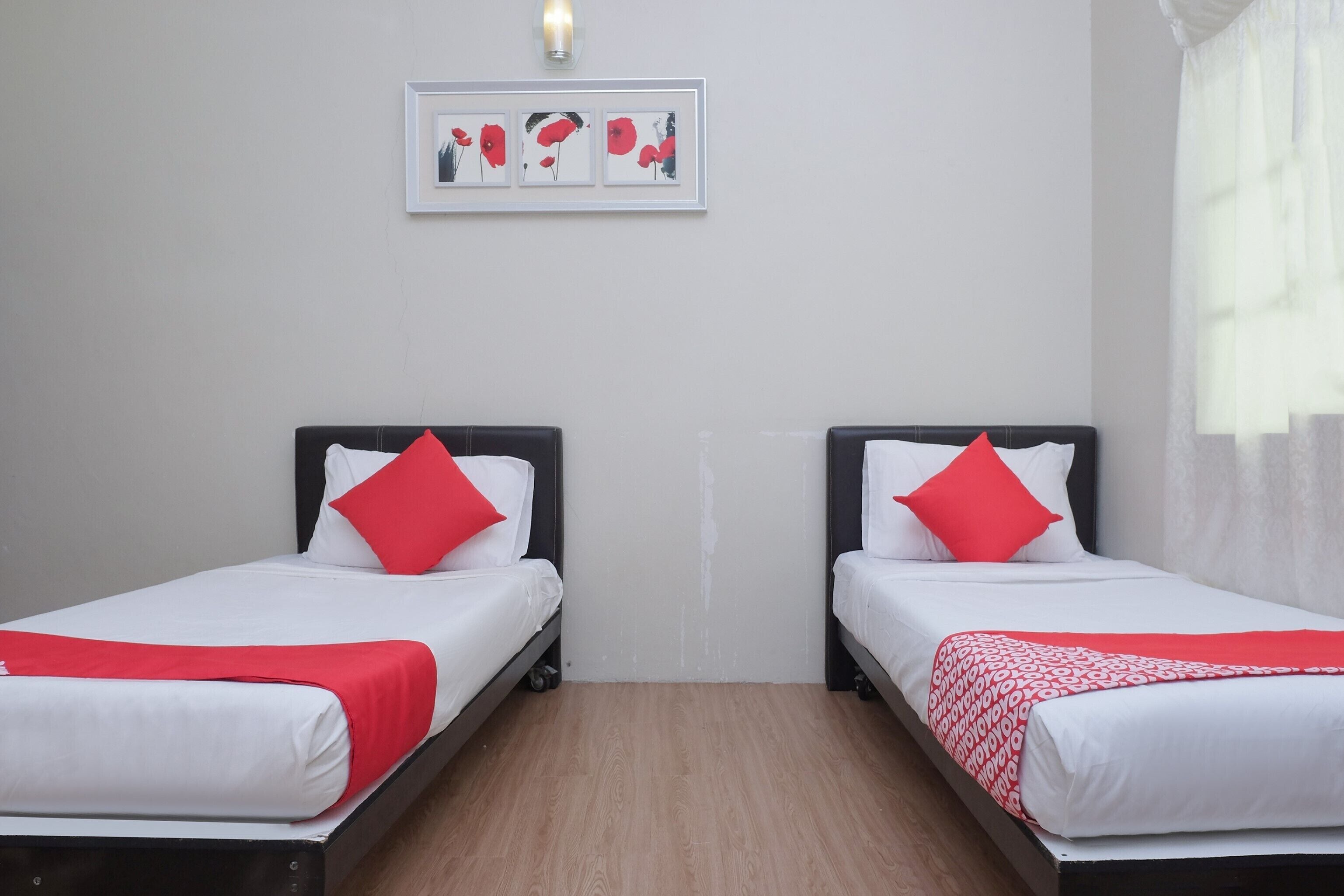 A hotel room with two single beds, each with white bedding and red pillows, a framed artwork above the beds, and a wooden floor.