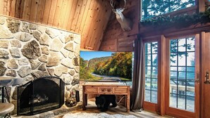 A cabin interior with a stone wall, a fireplace, a bear head decoration, a wooden table, and a scenic view through the glass door.