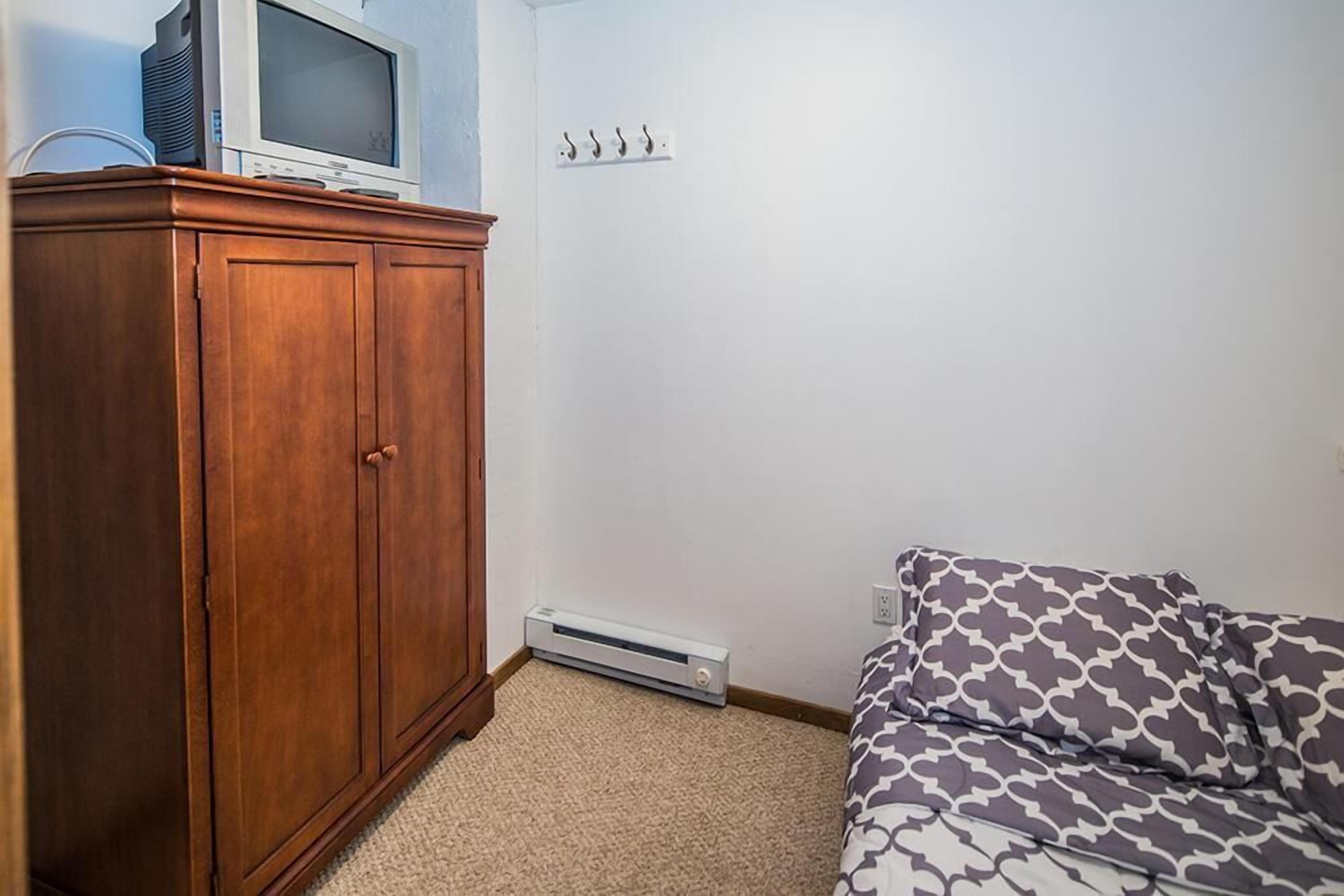 A bedroom with a wooden wardrobe, a bed with patterned bedding, a wall-mounted heater, and a television on top of the wardrobe.