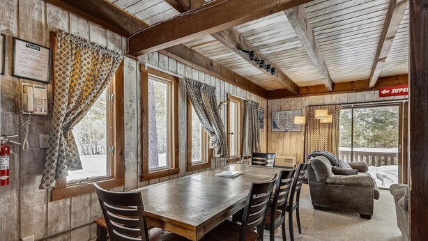 Interior acogedor de una cabaña con una mesa de comedor de madera, sillas, un sofá y ventanas grandes con cortinas.