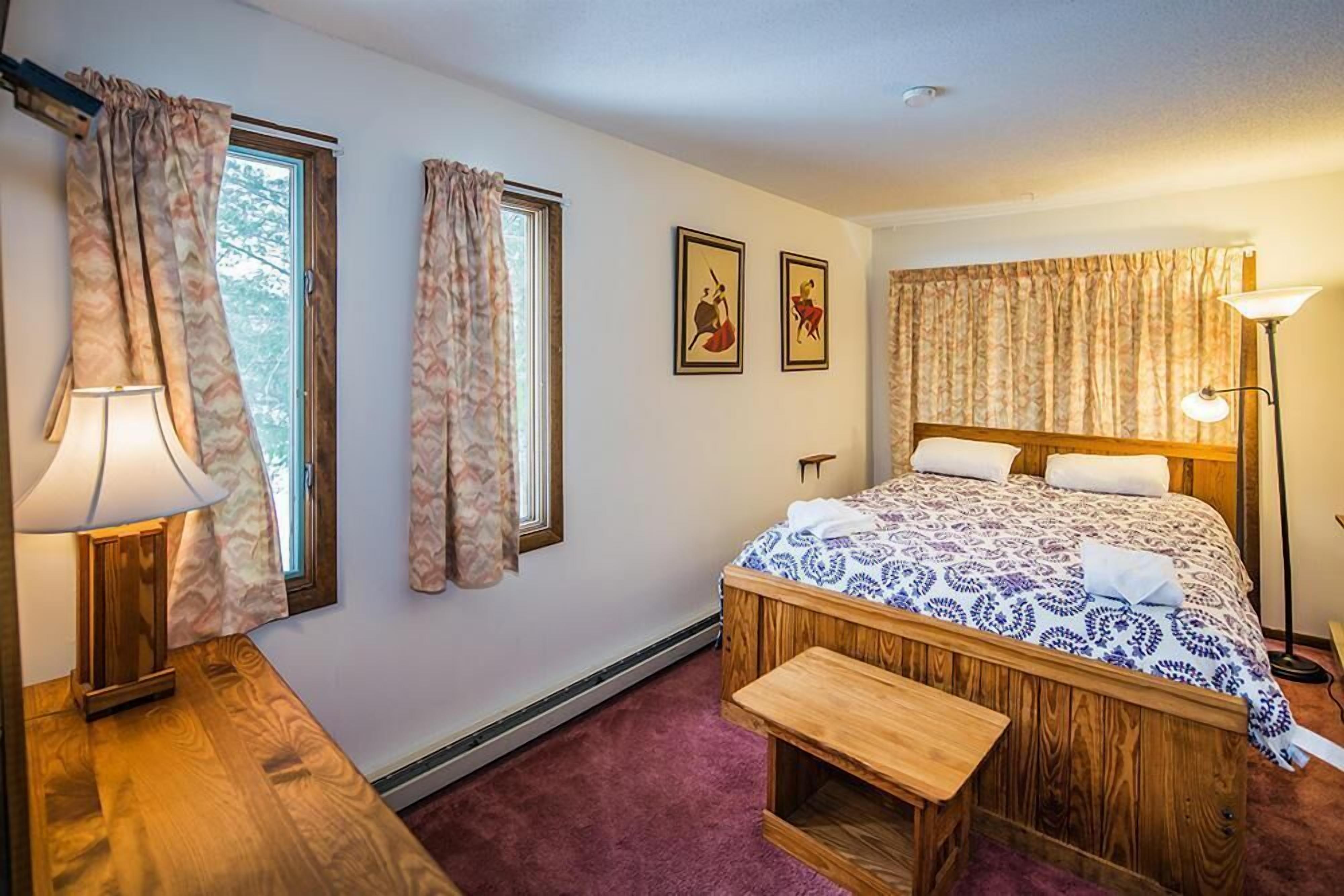 Une chambre avec un lit en bois, un banc, deux fenêtres avec des rideaux, une lampe et deux tableaux encadrés accrochés au mur.