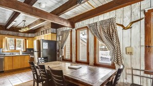 Interior rústico de una cabaña con una mesa de comedor de madera, sillas, una zona de cocina y ventanas amplias con cortinas.